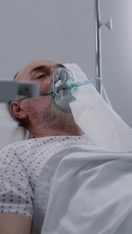 Vertical-shot-man-in-oxygen-mask-in-bed-in-hospital-ward.-Old-patient-with-pneumonia-during-lung-ventilation.-Modern-emergency-room-in-clinic.-Intensive-care-coronavirus-department-in-medical-facility.